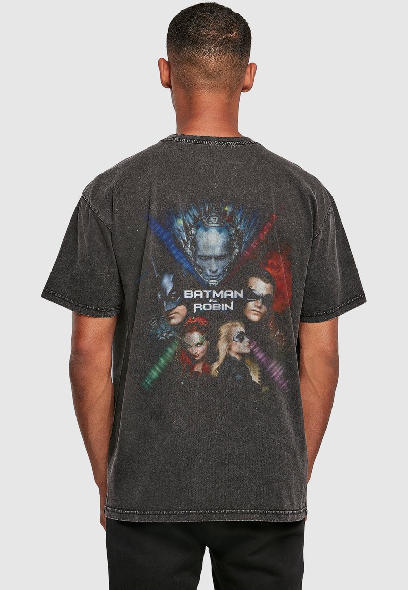 Homme portant un t-shirt noir délavé avec des personnages colorés Batman & Robin et un logo imprimé dans le dos sur un fond uni.