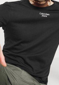 T-shirt noir à manches courtes en coton, avec un logo Calvin Klein Jeans blanc sur la poitrine. Texture lisse avec une coupe classique.