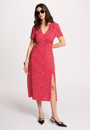 Mujer con vestido midi rojo con lunares blancos, escote en V, mangas cortas, abertura frontal, tacones negros, gafas de sol y pulsera dorada.