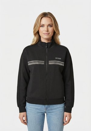 Femme aux cheveux blonds portant une veste noire zippée avec des rayures blanches à pois et le logo "GUESS", associée à un jean bleu clair.