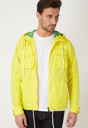 Hombre con chaqueta con capucha amarilla brillante sobre camisa blanca y jeans oscuros, mirando hacia un lado contra un fondo liso.