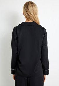 Zwarte shirt met lange mouwen met een gladde structuur, klassieke kraag en witte piping accenten op de manchetten. Eenvoudig ontwerp zonder zichtbare sluitingen.