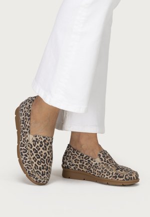 Feet wearing leopard print slip-on loafers with tan soles, paired with white cropped wide-leg pants against a white background.