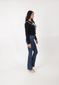 Suéter negro con detalles de cuello blanco, combinado con jeans acampanados ajustados en azul y sandalias blancas de tacón, de pie contra un fondo blanco.
