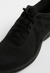 Svart atletisk sko med ett texturerat meshuppdrag, rund tå, platta snören och minimalistisk design; har en vadderad sula och logodetalj.