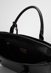Sac à main en cuir noir à texture lisse, équipé de deux poignées et d'une poche intérieure zippée. Comprend une étiquette logo à l’intérieur et un accent doré sur la poignée.