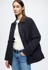 Námornícky ľahký kabát s hnedým semišovým golierom, vybavený patentkami a zipsom, kombinovaný s bielym rebrovaným topom a modrými džínami.