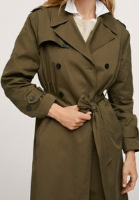 Olivengrøn trenchcoat med dobbeltbrystet design, sorte knapper, bælte i taljen og skulderklapper. Glat stof og skræddersyet pasform.