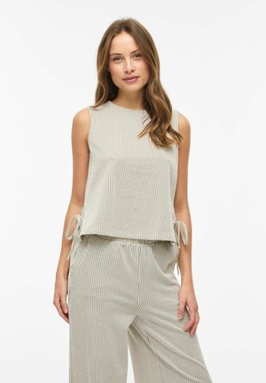 Mujer con top sin mangas de rayas verticales beige y blancas y pantalones holgados a juego con lazos laterales, posando sobre un fondo blanco liso.