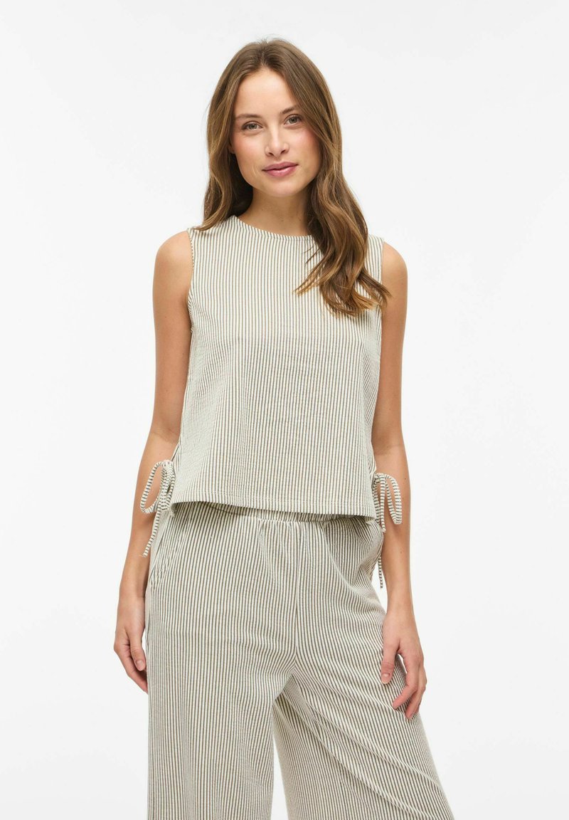 Femme portant un haut sans manches à rayures verticales beige et blanches ainsi qu'un pantalon ample assorti avec des liens sur les côtés, debout devant un fond blanc uni.