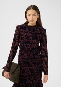 Langärmliges, tailliertes Kleid in Bordeauxrot mit schwarzem Blumenmuster, strukturiertem Stoff, gerafftem Detail und rüschengem Saum. Als Accessoire wird eine grüne Clutch mitgeführt.