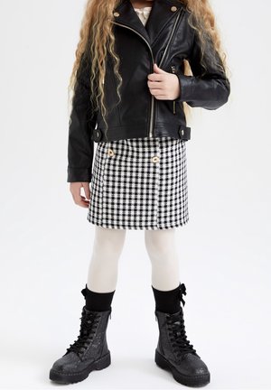 Bambino che indossa una giacca di pelle nera, una gonna a quadri bianca e nera con bottoni, collant bianchi e stivali da combattimento neri scintillanti.
