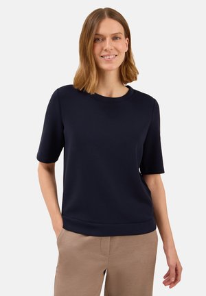 Donna che indossa una maglietta blu scuro a maniche corte e pantaloni beige, in piedi con una mano in tasca su uno sfondo bianco uniforme.