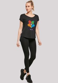 Svart kortermet t-skjorte med et flerfarget Hogwarts-omslagdesign, brukt med svarte leggings og svarte joggesko.