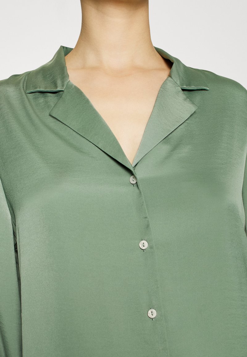 Blusa verde in satin con colletto a pipistrello, dotata di sei piccoli bottoni sul davanti, dalla texture liscia e una vestibilità leggermente rilassata.