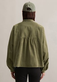 Veste en velours côtelé vert olive avec des manches bouffantes et un dos froncé, associée à une casquette verte assortie avec une sangle réglable.