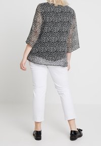 Femme portant un blouse à pois noir et blanc avec des manches transparents, un pantalon blanc court et des mocassins à pompons noirs, se tenant de dos.