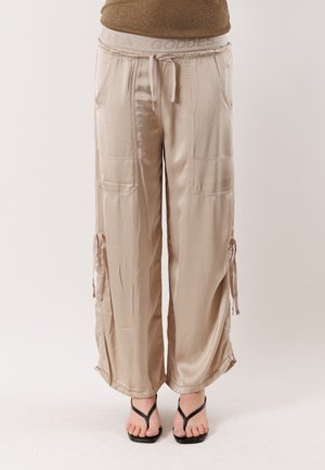 Pantaloni beige larghi a gamba larga con tasche applicate, vita con coulisse, fiocchi laterali, indossati con sandali neri a punta quadrata.