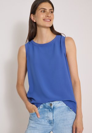 Femme souriante regardant vers la droite, portant un haut bleu sans manches et un jean en denim bleu clair, une main dans la poche.