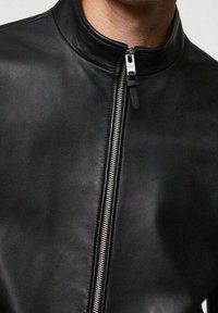 Uomo che indossa una giacca in pelle nera con una cerniera verticale argentata e un colletto corto in piedi visibile.