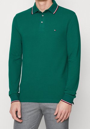 Homme portant un polo à manches longues vert avec un col et des poignets rayés rouges et blancs, associé à un pantalon gris à motifs.