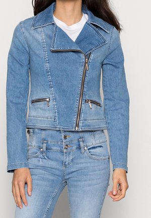 Frau trägt eine hellblaue Jeansjacke mit asymmetrischem Reißverschluss und zwei Reißverschlusstaschen, kombiniert mit hoch taillierten Jeans.