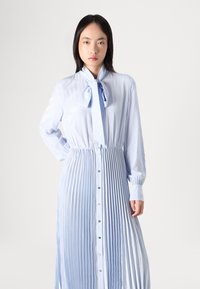 Vestido plissado azul claro com gola amarrada, mangas longas e fechos de botões na frente. Textura suave e design fluido.