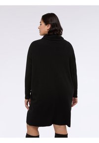 Abito maglione nero oversize con collo a scialle, maniche lunghe e tessuto morbido, che arriva a metà coscia. Design semplice, senza motivi o accenti.