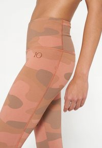 Leggings a vita alta in un motivo camouflage rosa e marrone, realizzati in tessuto morbido e elastico con dettagli cuciti e il numero "10" sulla fascia.