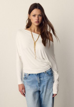 Ženska v beli dolgoročni majici z golimi rameni, ohlapnih svetlo modrih kavbojkah in dolgi zlati večplastni verižici, stoji pred enobarvnim ozadjem.