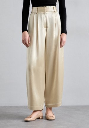 Persona che indossa pantaloni beige in raso a vita alta e gamba larga con pieghe, top nero a maniche lunghe e scarpe basse color nude, in piedi su pavimento grigio.