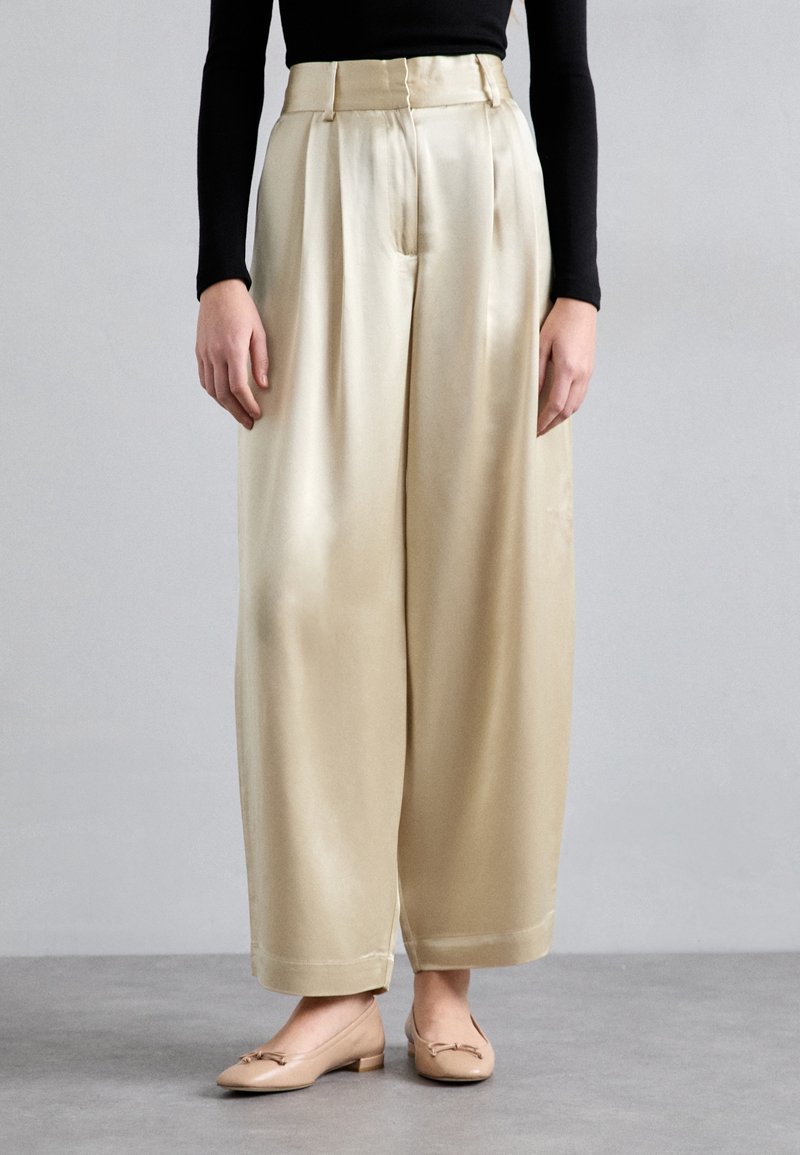 Personne portant un pantalon large en satin beige taille haute avec des plis, un haut noir à manches longues et des chaussures plates couleur nude, debout sur un sol gris.