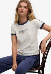 Wit T-shirt met marineblauwe accenten en "SUPERDRY" opdruk aan de voorkant, gecombineerd met donkerblauwe sweatpants met een gedrukt nummer.