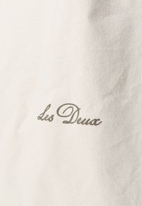 Λευκό βαμβακερό ύφασμα με κεντημένο το "Les Deux" σε κυρτή, ανοιχτό γκρι γραφή, με υφές που διαθέτει μια λεπτή κοκκώδη διακριτική επιφάνεια.