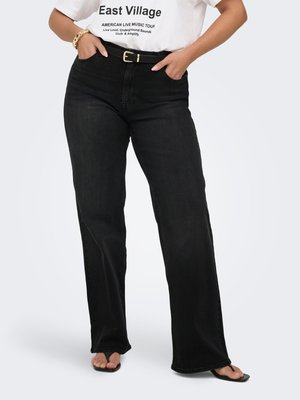Person iført sorte jeans med vide ben og sort bælte, en hvid t-shirt med tryk "East Village" og sorte sandaler med åbne tæer.