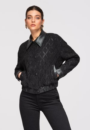 Kvinde med mørkt hår iført en sort tekstureret jakke med læderkrave og manchetter, hænderne i lommerne, iført sorte bukser, neutral baggrund.