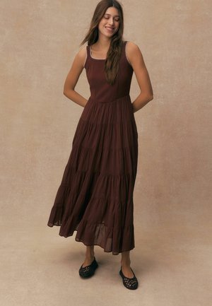 Woman with long brown hair wearing a sleeveless, tiered brown maxi dress and black woven flats, standing against a beige backdrop.