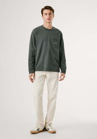 Camisa de algodón de manga larga verde oscuro con un bolsillo en el pecho, combinada con pantalones color crema y zapatillas color blanco roto.