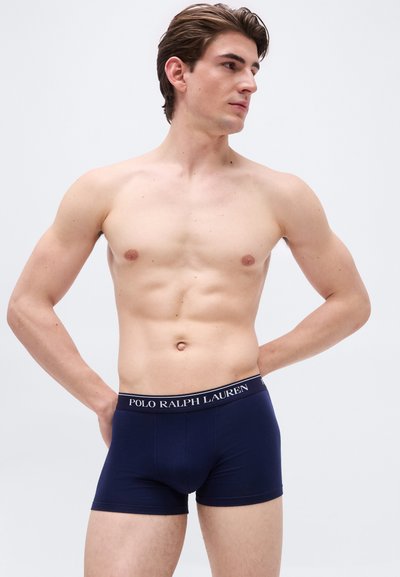 Νεαρός άντρας με κοντά καστανά μαλλιά ποζάρει χωρίς πουκάμισο, φορώντας σκούρα μπλε boxer Ralph Lauren Polo, κοιτάζοντας προς τα δεξιά του με τα χέρια πίσω από την πλάτη.