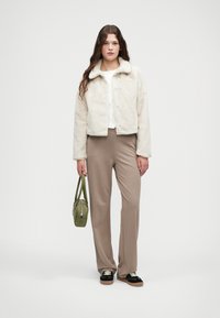 Lažni krzneni kaput u boji zemlje s ovratnikom, s kopčanjem sprijeda, uparen s bež hlačama visokog struka i malom zelenom torbicom.