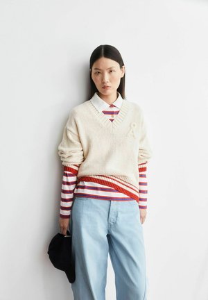 Jonge vrouw met steil zwart haar draagt een crème-kleurige gebreide trui over een roodgestreept overhemd, lichtblauwe spijkerbroek, en houdt een zwarte pet vast tegen een witte achtergrond.