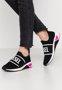 Mustat liuku-ukot kengät, joissa valkoinen rohkea "DIESEL"-logo, joustava päällinen, teksturoitu pohja ja vaaleanpunainen yksityiskohta kantapäässä. Yhdistetty revenneisiin mustiin farkkuihin.