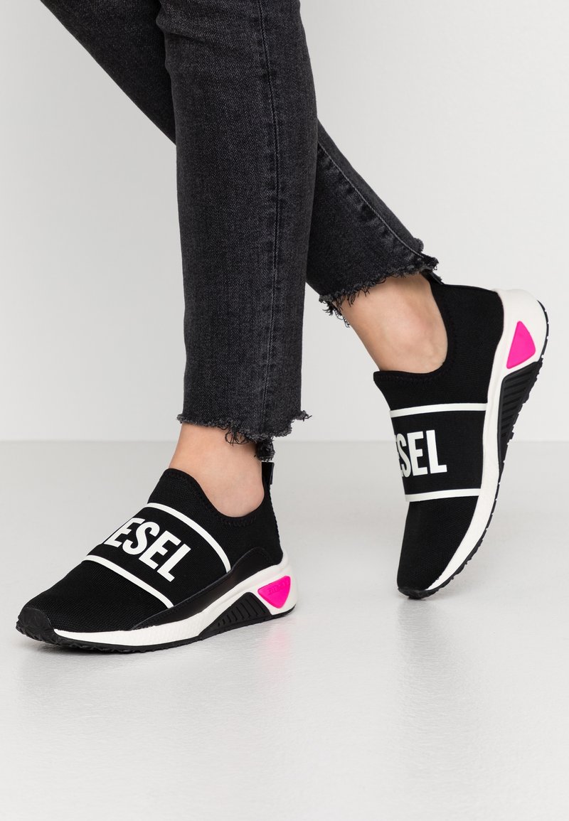 Mustat liuku-ukot kengät, joissa valkoinen rohkea "DIESEL"-logo, joustava päällinen, teksturoitu pohja ja vaaleanpunainen yksityiskohta kantapäässä. Yhdistetty revenneisiin mustiin farkkuihin.