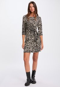 Leopardenmusterkleid mit Dreiviertelärmeln, taillierter Taille und einer gebundenen Detail in Schwarz und Beige. Getragen mit schwarzen Stiefeletten.