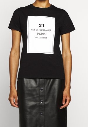 Osoba nosící černé tričko s bílým čtvercovým potiskem s nápisem "21 Rue St-Guillaume Paris Karl Lagerfeld" a černou koženou sukní na knoflíky.