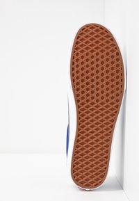 Kengän pohja ruskealla kumilla waffle-kuviolla, valkoinen välikerros ja sininen kangaspinta, asetettu pystysuoraan valkoista kulmaa vasten.