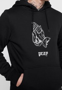 Černá mikina s kapucí s bílou grafikou modlících se rukou držících růženec nad slovem "modlit" ve gotickém písmu, kterou nosí osoba s tetováním.