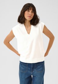 Top en maille sans manches blanc avec un col en V, un ourlet côtelé et une texture douce ; associé à un jean en denim bleu avec des poches arrière.