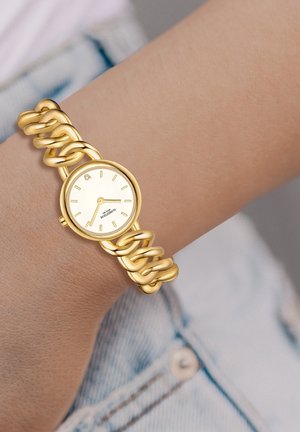 Montre-bracelet en maillon doré avec un cadran rond blanc et des marqueurs d'heure minimalistes, portée sur un poignet au-dessus d'un tissu en denim bleu clair.