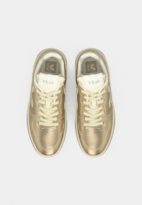 Χρυσά αθλητικά παπούτσια από βίγκαν δέρμα με διάτρητες λεπτομέρειες, εκρού κορδόνια και επενδυμένες γλώσσες με την επωνυμία "VEJA" στο επάνω μέρος.